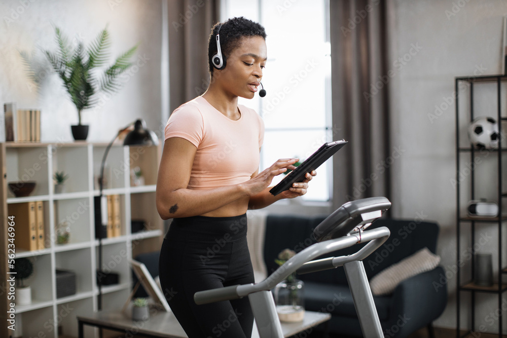 Close up of young active african businesswoman in sportswear and headset training at home working with tablet, doing cardio exercise on treadmill. Concept of sport, business. Close up of young active african businesswoman in sportswear and headset training at home working with tablet, doing cardio exercise on treadmill. Concept of sport, business.