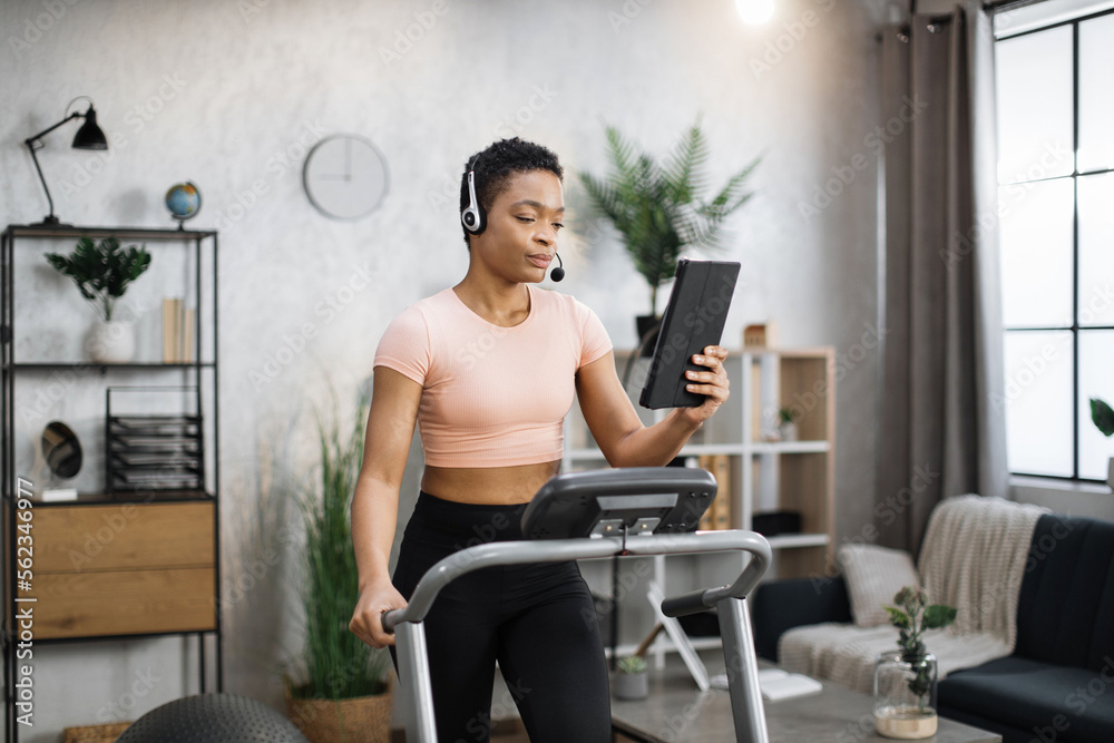 Fototapeta premium Close up of young active african businesswoman in sportswear and headset training at home working with tablet, doing cardio exercise on treadmill. Concept of sport, business.