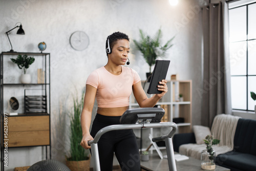 Close up of young active african businesswoman in sportswear and headset training at home working with tablet  doing cardio exercise on treadmill. Concept of sport  business.