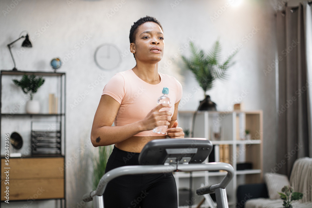 Attractive young sports african american woman holding bottle with water working out, running, doing cardio training on treadmill, indoor on background of modern apartment.