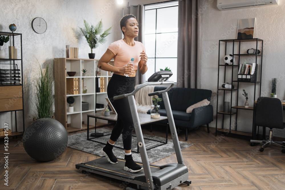 Fototapeta premium Portrait of active and dynamic young charming thirsty african woman holding water bottle doing sport fitness at home running on treadmill indoor. Tone your body. Perfect shape. Improving endurance.