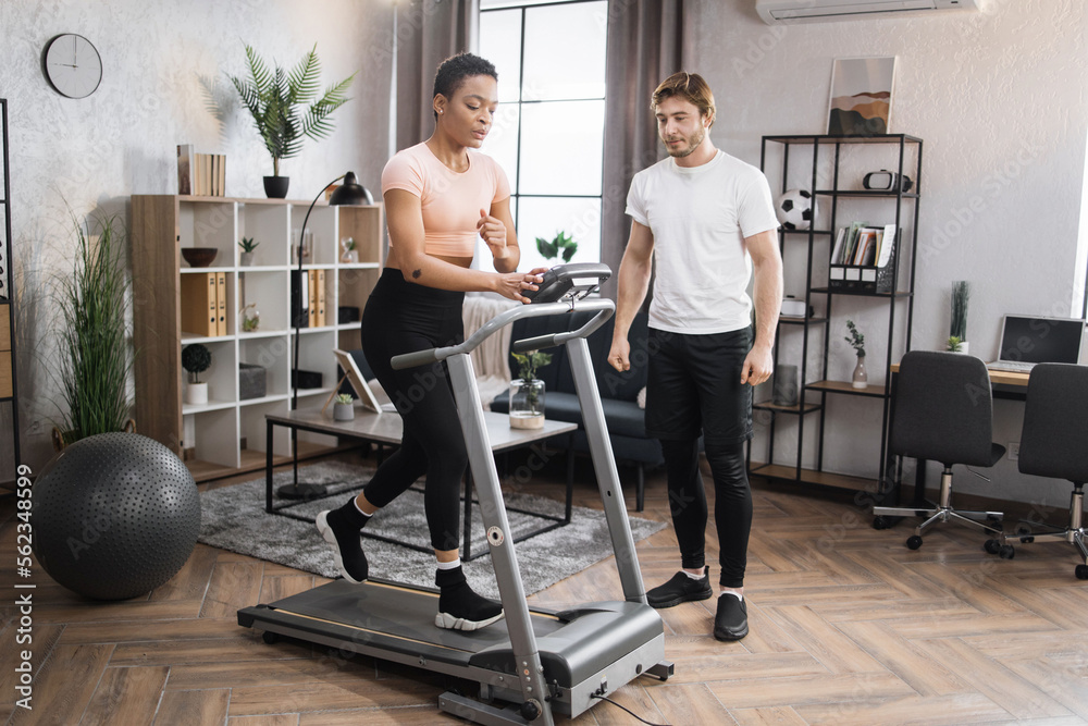 Fototapeta premium Portrait of focused young african female wearing sportswear and using treadmill and her trainer bearded caucasian male. Home fitness workout sporty woman and man training on running track.