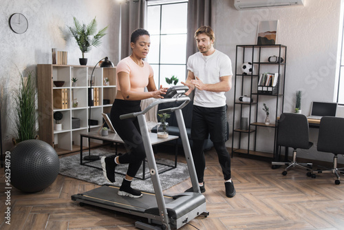 Portrait of focused young african female wearing sportswear and using treadmill and her trainer bearded caucasian male. Home fitness workout sporty woman and man training on running track.