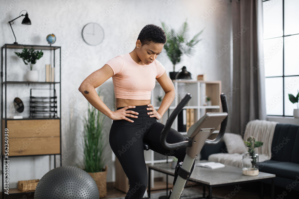 Fototapeta premium Young attractive sporty tired african american woman in sportswear having morning workout at modern light apartment. Healthy athletic female doing cardio on stationary bike.