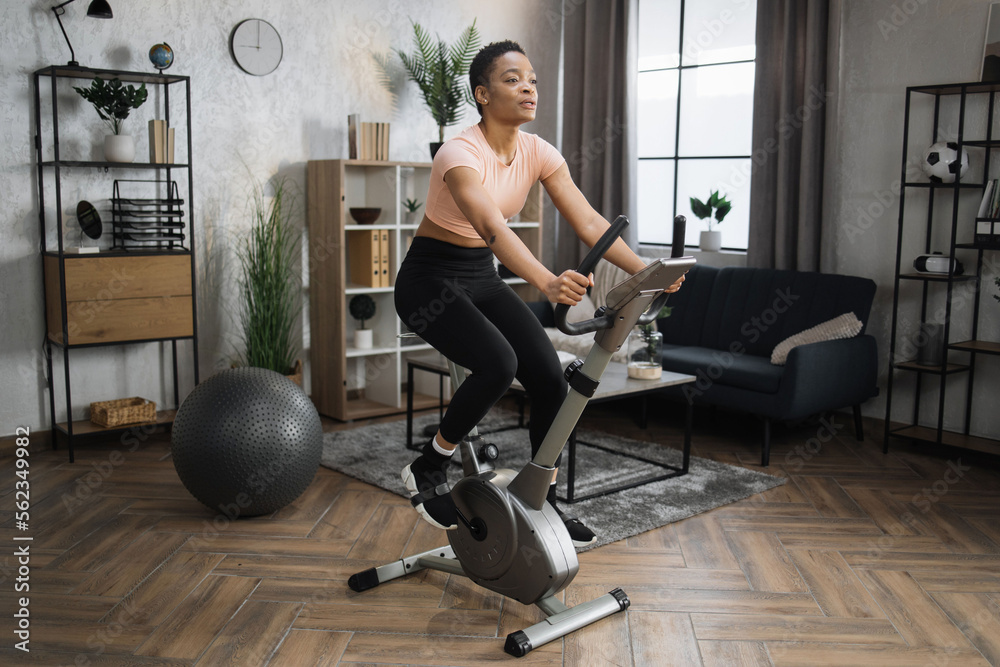 Full length portrait of young sportswoman training at home gym doing cardio on stationary bike. Cardio training, exercising legs, cardio workout indoors. Full length portrait of young sportswoman training at home gym doing cardio on stationary bike. Cardio training, exercising legs, cardio workout indoors.