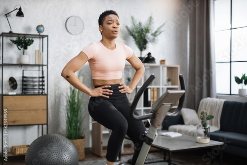 Young attractive sporty tired african american woman in sportswear having morning workout at modern light apartment. Healthy athletic female doing cardio on stationary bike.