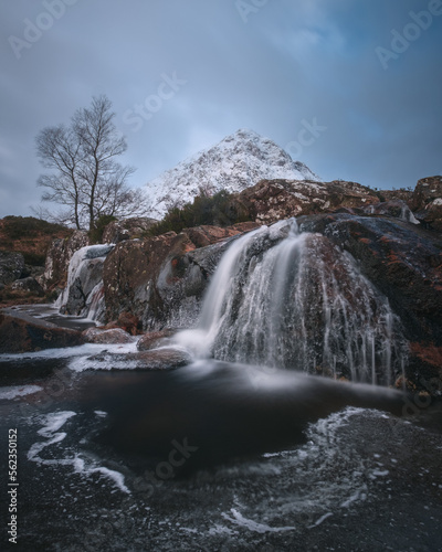 Mountain river and partially frozen waterfall at the foot of the snow-covered Buachaille Etive Mor. Etive Mor Waterfall. At the entrance to the valley of Glencoe in the Scottish Highlands, Scotland