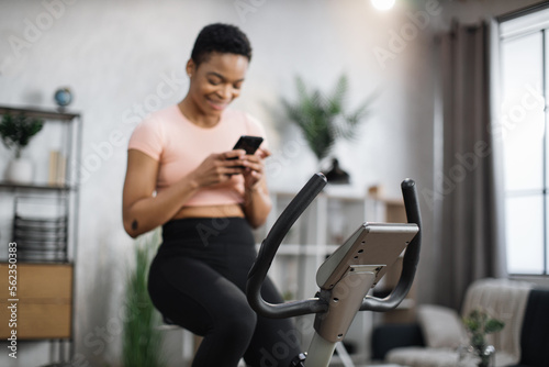 Focus on screen connected online to live streaming subscription service for biking exercise. Low angle view of african female cyclist, exercising at home, riding stationary bike simulator.