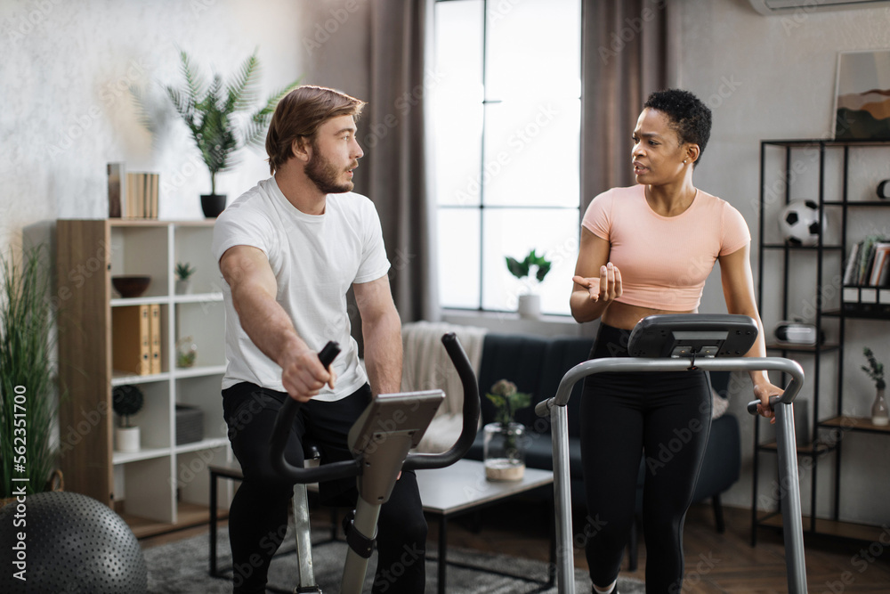 Fototapeta premium Young couple doing cardio on stationary bike and treadmill. Attractive female and male working out at home. Woman running on treadmill and man training on exercise bike.
