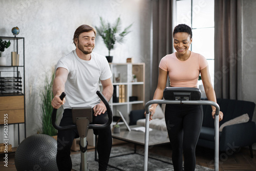 Young couple doing cardio on stationary bike and treadmill. Attractive female and male working out at home. Woman running on treadmill and man training on exercise bike.