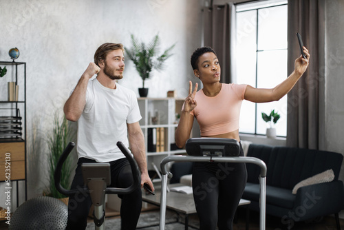 African american sports woman in sportswear running on treadmill and fit caucasian man cycling bike and making selfie or having online video call.