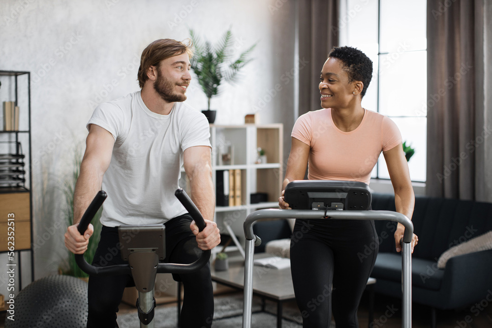Home fitness workout sporty people training on exercise machines indoors. Portrait of focused caucasian male and african female wearing sportswear using exercise bike and treadmill. Home fitness workout sporty people training on exercise machines indoors. Portrait of focused caucasian male and african female wearing sportswear using exercise bike and treadmill.