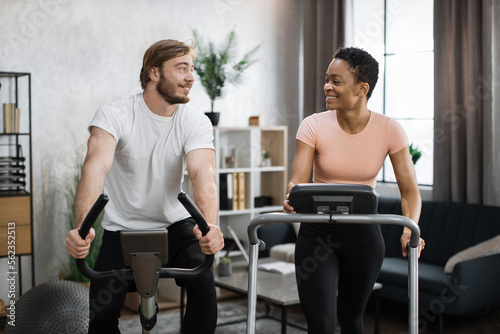 Home fitness workout sporty people training on exercise machines indoors. Portrait of focused caucasian male and african female wearing sportswear using exercise bike and treadmill.
