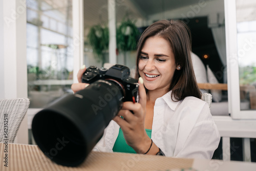 Adorable female young brunette model hods camera watching photos on screen, smiling satisfied by shooting results. Attractive young American woman at cafe flipping through photos. 