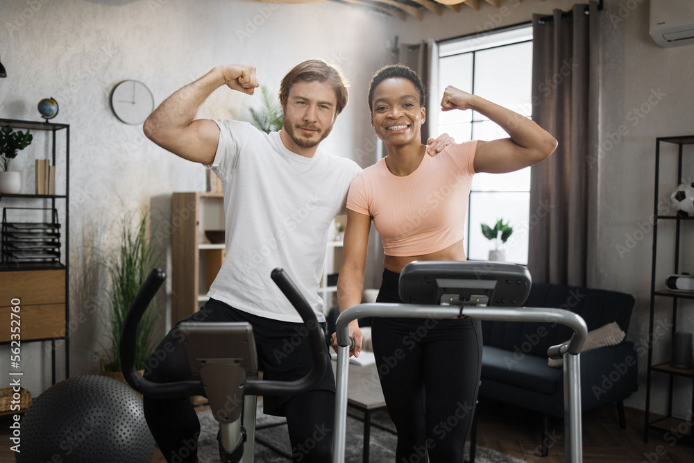 Obraz premium Smiling african woman running on treadmill and caucasian man training on exercise bike showing biceps looking at camera. Young couple doing cardio on stationary bike and treadmill.