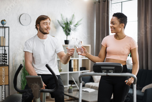 Portrait of young couple doing cardio on stationary bike and treadmill clinking bottles of water  cheers happy to finish training. Attractive female and male working out on fitness.