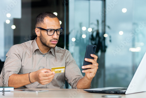 Dissatisfied and angry businessman in office trying to make purchase in online store, hispanic man holding bank credit card and smartphone in hands, man cheated received error fraud. photo