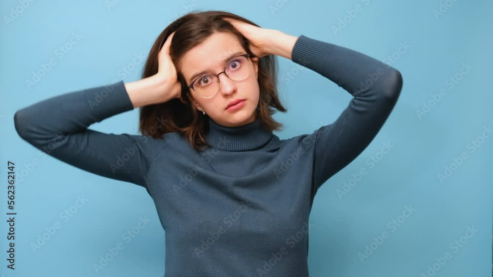 Video portrait of a stressed student girl with glasses and a sweater holding her head on a blue background, 4K. Horrible, stress, shock. Emotions, facial expression Video portrait of a stressed student girl with glasses and a sweater holding her head on a blue background, 4K. Horrible, stress, shock. Emotions, facial expression
