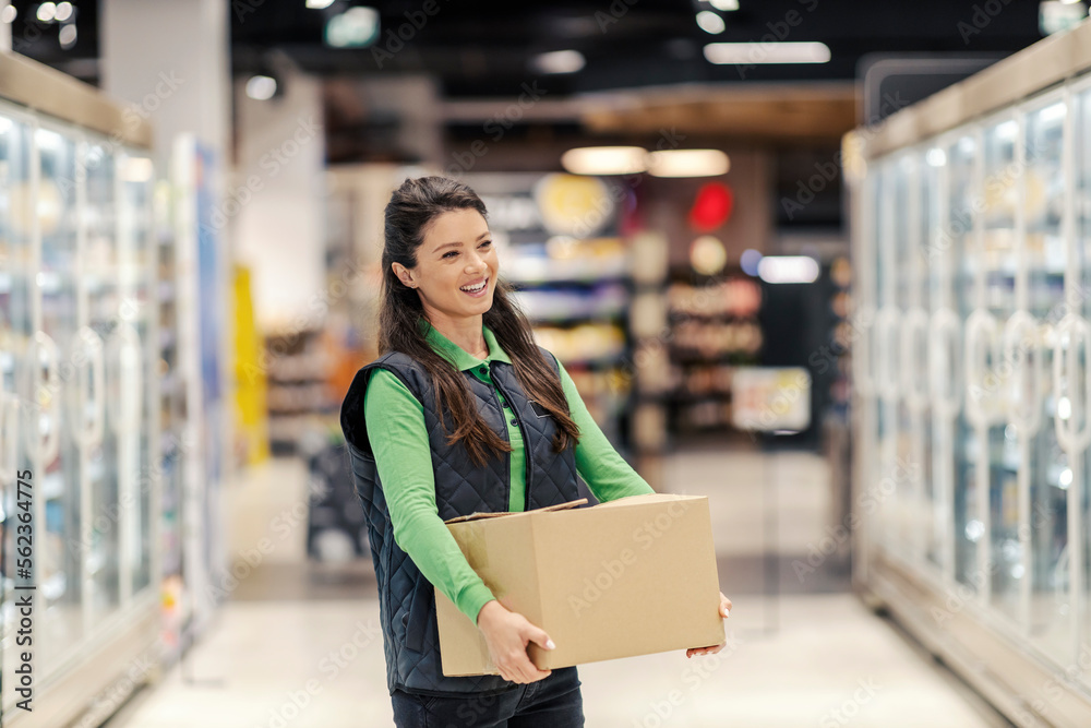 A happy stock clerk is carrying box with groceries in supermarket. A happy stock clerk is carrying box with groceries in supermarket.
