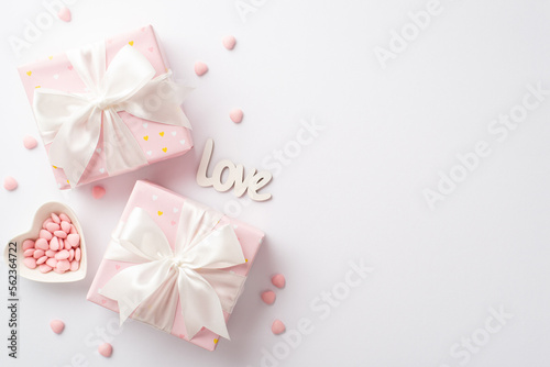 Valentine's Day concept. Top view photo of pastel pink gift boxes with silk ribbon bows inscription love and heart shaped saucer with sprinkles on isolated white background with copyspace