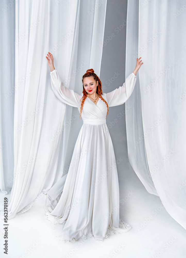 beautiful red-head woman in white long dress with long curly hair holding posing in white studio with curtains beautiful red-head woman in white long dress with long curly hair holding posing in white studio with curtains