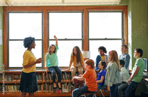 Education, learning and student with questions for teacher in middle school classroom. Library, scholarship group and girl learner raising hand to answer question, studying or help with black woman. photo