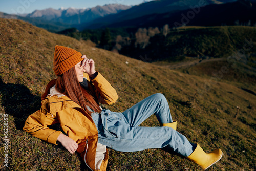 Woman beautifully lying on a hill on the grass rest smile with teeth looking at the mountains in the autumn in a yellow raincoat and jeans happy sunset trip on a hike, freedom lifestyle 