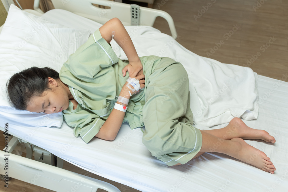 Obraz premium Asian young beautiful female lying on the bad having stomach ache and IV saline drip to back of hand lying on bed in hospital, medical and health concepts.