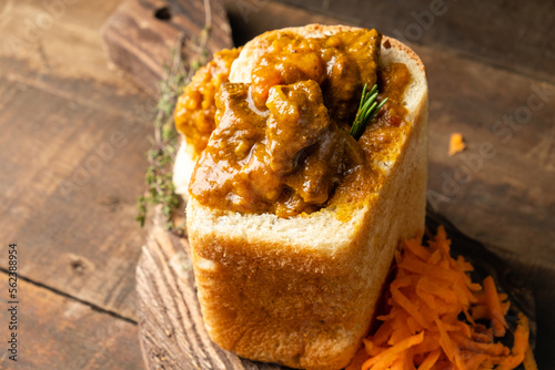 Bunny chow curry with meat and vegetables served in white bread and carrot salad on a wooden background, close-up. photo