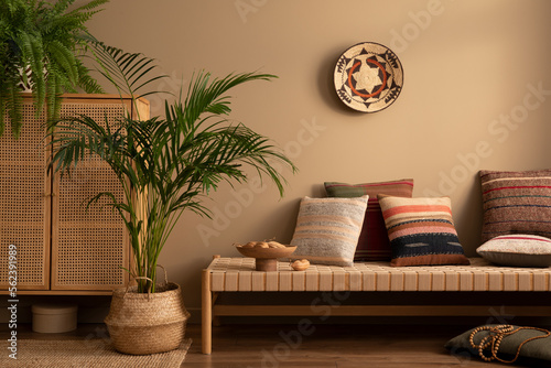 Warm and cozy ethno living room interior with couch, patterned pillows, plants i flowerpots, fern, rattan sideboard, basket on wall, wooden bowl and personal accessories. Home decor. Template. photo