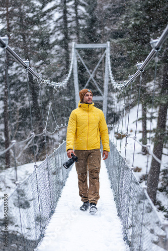 Young caucasian man walking on a hanging bridge on a snowy day holding a camera in winter clothes.