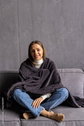 Cute young woman relaxing o the sofa wrapped in a soft grey blanket at home