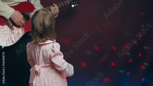 Little girl on stage in vintage dress, she sings into microphone and dances, her father plays acoustic guitar. Color music is shining. Performance on stage. Early development of children