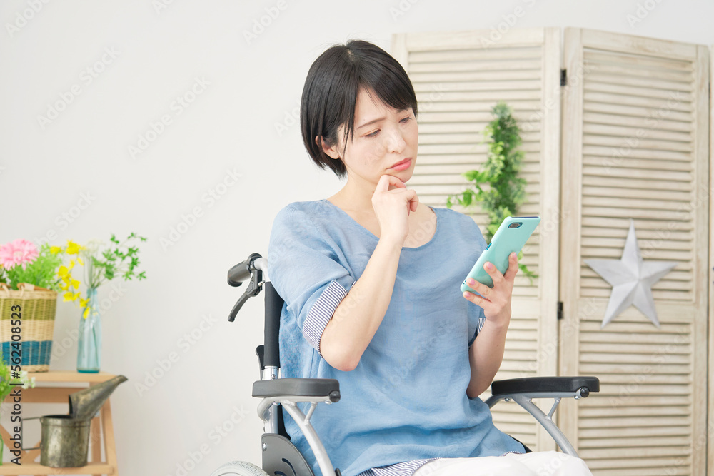 家でスマホを持って考える車椅子に乗った障がい者女性 家でスマホを持って考える車椅子に乗った障がい者女性