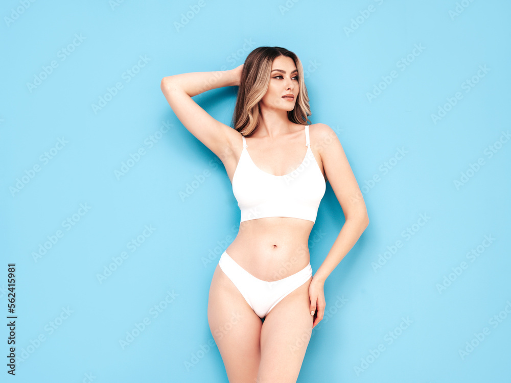 Fashion portrait of young beautiful woman. Attractive carefree  blond model wearing pure white lingerie. Hot tanned confident blonde posing near blue wall in studio, showing sexy natural body