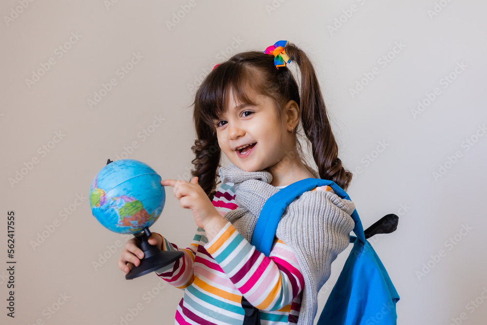 a happy little preschool girl with a backpack and a globe on a light background, space for text. Preschool education, traveling with children a happy little preschool girl with a backpack and a globe on a light background, space for text. Preschool education, traveling with children