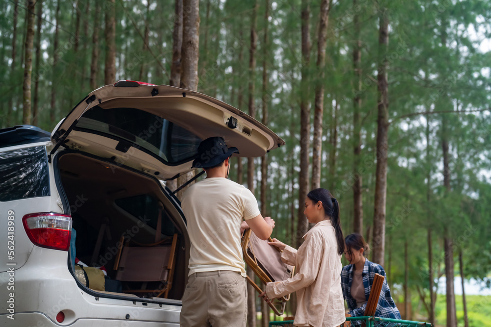 Group of Asian people friends enjoy outdoor lifestyle road trip and camping together on summer holiday travel vacation. Man and woman taking off camping supplies from car trunk at natural park.