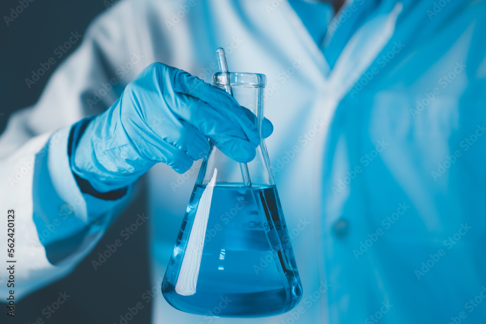 Scientist hand hold erlenmeyer flask with stirring rod filled with blue sample chemicals in chemistry science laboratory. Glassware in medical research. Scientific lab for Biotechnology.
