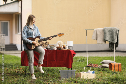 Woman holding guitar near table with different items on garage sale in yard