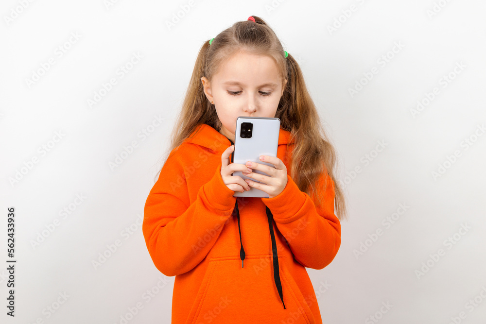 A little girl in an orange suit looks at her phone. Dependence of a generation of children on gadgets. On a white background. A little girl in an orange suit looks at her phone. Dependence of a generation of children on gadgets. On a white background.