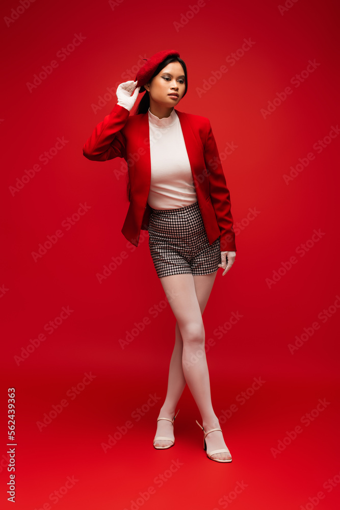 Asian woman in gloves touching beret and posing on red background Asian woman in gloves touching beret and posing on red background