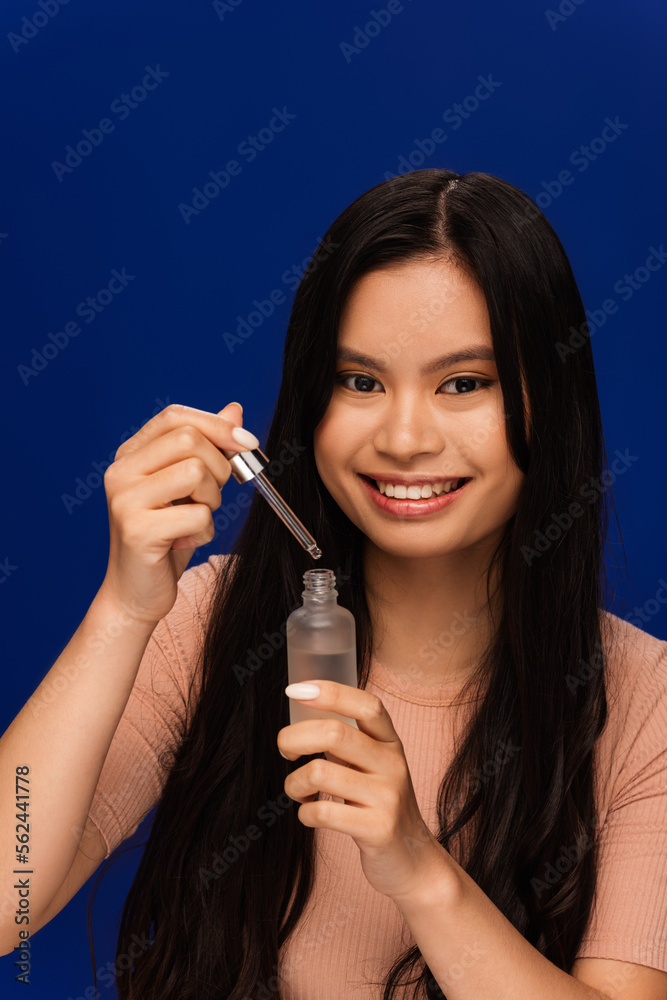 Smiling asian woman holding cosmetic serum and dropper isolated on blue Smiling asian woman holding cosmetic serum and dropper isolated on blue