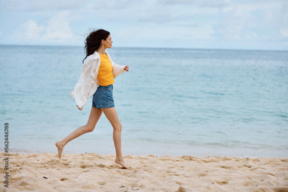 Sports woman runs along the beach in summer clothes on the sand in a yellow tank top and denim shorts white shirt flying hair ocean view, beach vacation and travel Sports woman runs along the beach in summer clothes on the sand in a yellow tank top and denim shorts white shirt flying hair ocean view, beach vacation and travel