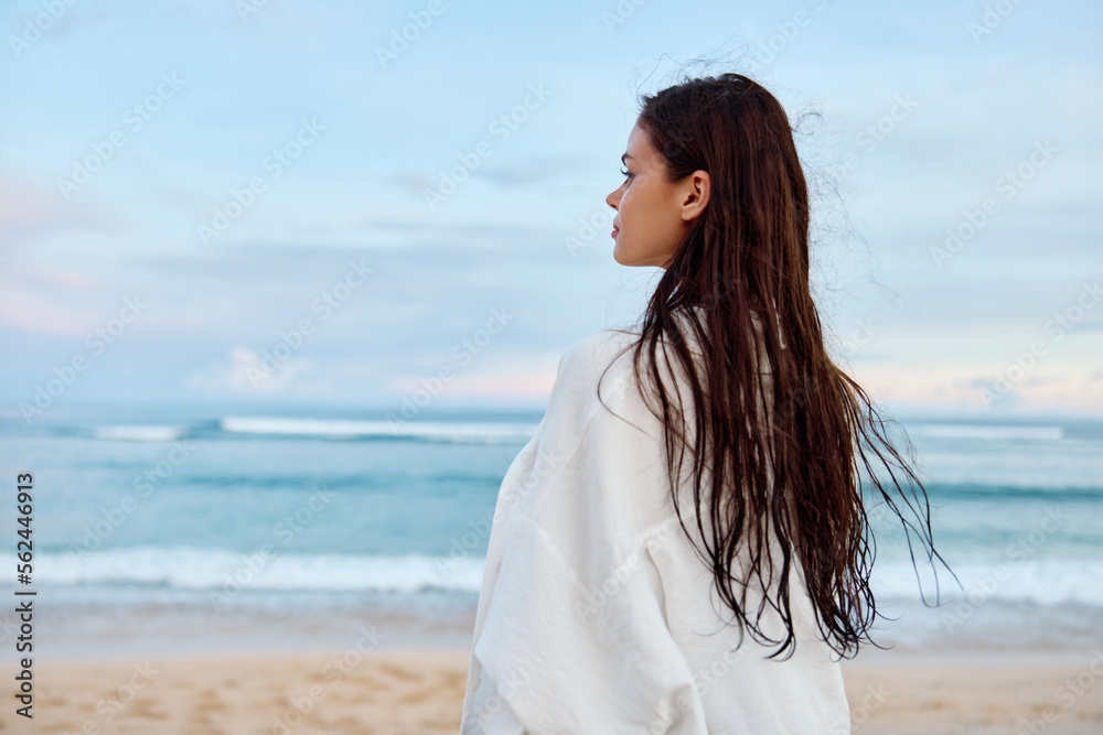 Happy tanned slender woman in white swimsuit shirt and denim shorts walks on the beach with her back to the camera on the sand by the ocean with wet hair after swimming, sunset light Happy tanned slender woman in white swimsuit shirt and denim shorts walks on the beach with her back to the camera on the sand by the ocean with wet hair after swimming, sunset light