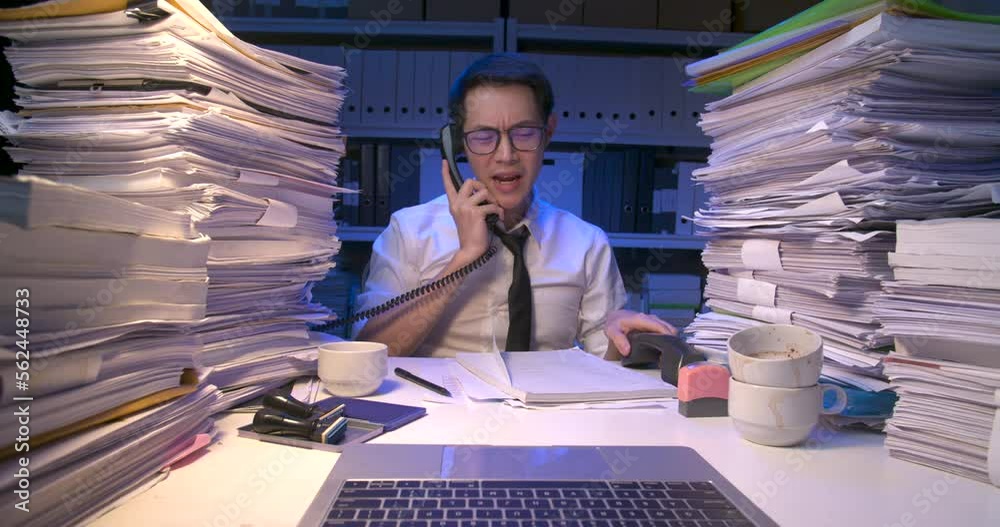 Busy asian business man is sitting at desk covered with stack of paperwork and having multiple answering phone calls. Alone male working multi tasking and holding many telephone at late night.