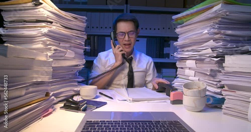 Busy asian business man is sitting at desk covered with stack of paperwork and having multiple answering phone calls. Alone male working multi tasking and holding many telephone at late night.