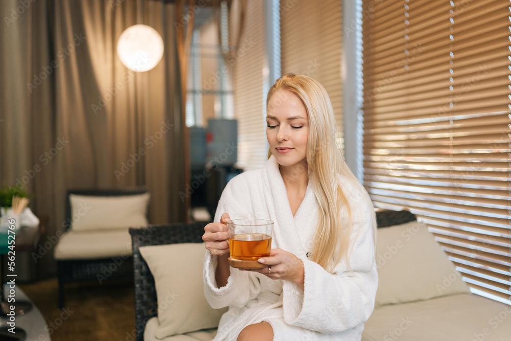 Portrait of gorgeous young woman in white bathrobe holding cup with fresh herbal tea, sitting on sofa by window in spa salon after treatment procedures. Concept of luxury professional massage.