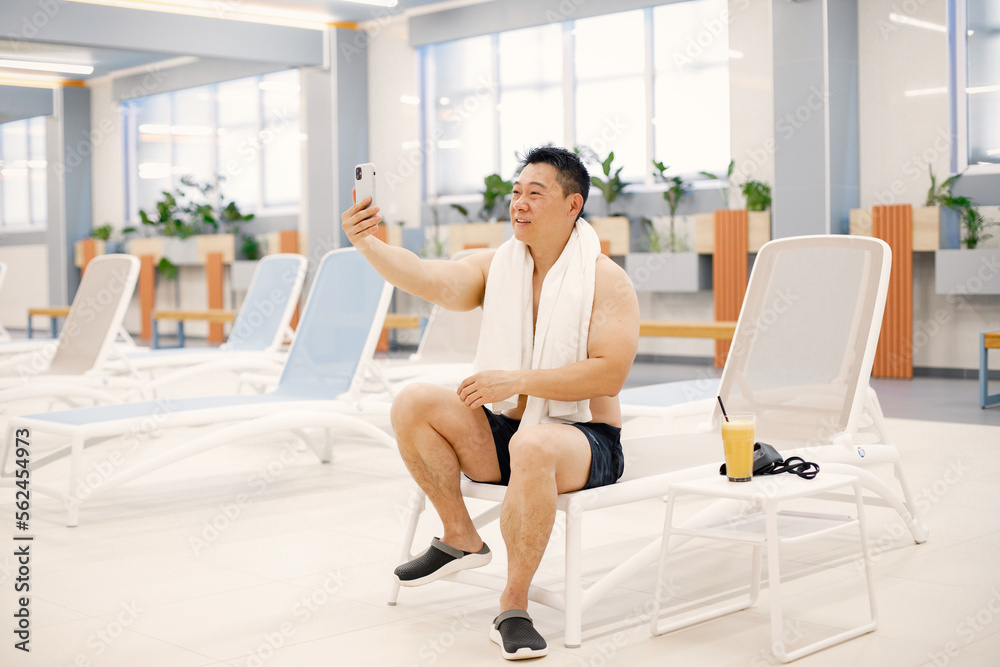Asian man sitting on a sunbed in indoors swimming pool and make a selfie Asian man sitting on a sunbed in indoors swimming pool and make a selfie