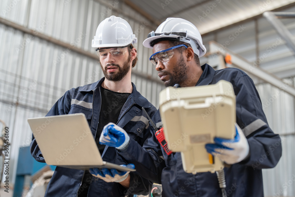 Male engineers wearing vest with helmet safety using labtop and remote controller control inspection robot arm welding machine in factory. Team of technician automation robot system. Male engineers wearing vest with helmet safety using labtop and remote controller control inspection robot arm welding machine in factory. Team of technician automation robot system.