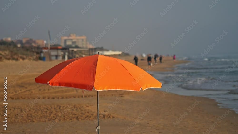 An orange beach umbrella against the background of a beach with people walking, shot in slow motion. Bar and buildings in the background. An orange beach umbrella against the background of a beach with people walking, shot in slow motion. Bar and buildings in the background.